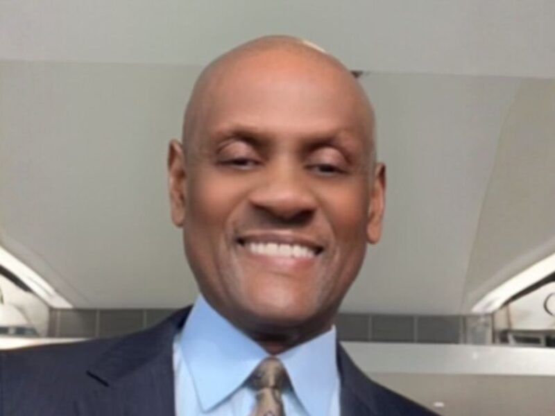 Smiling bald man wearing a suit, light blue shirt, and patterned tie indoors.