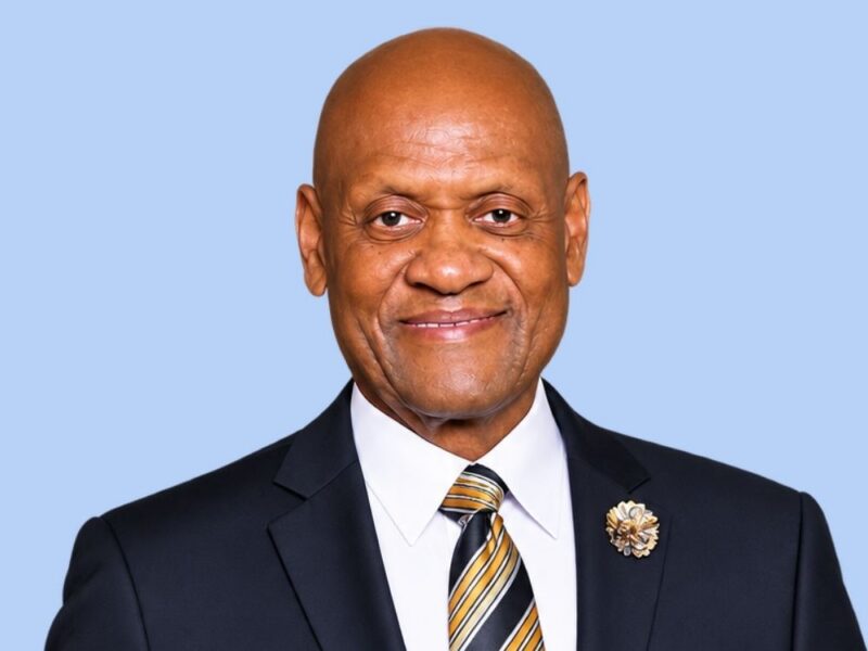 Portrait of a bald man in a suit and striped tie against a light blue background.