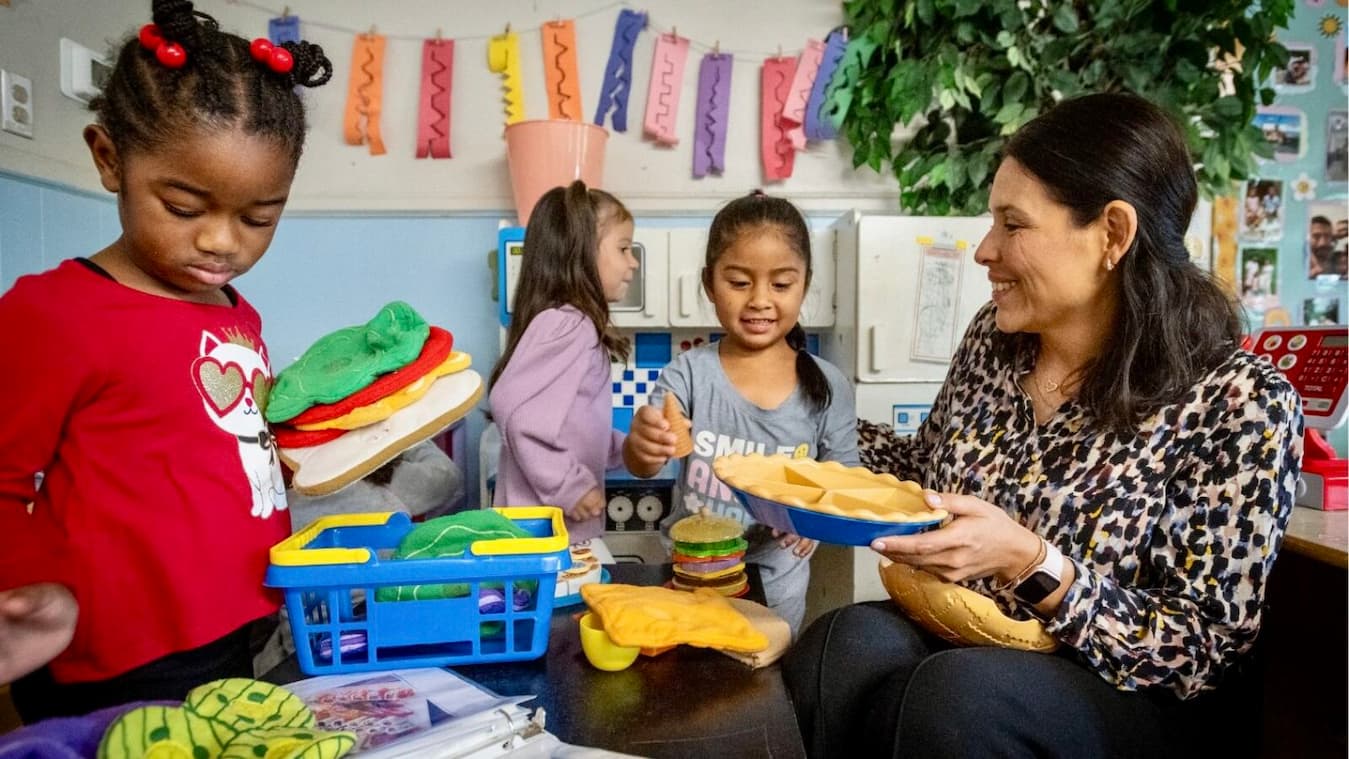 Teacher and three children playing with toy food in a colorful classroom setting.