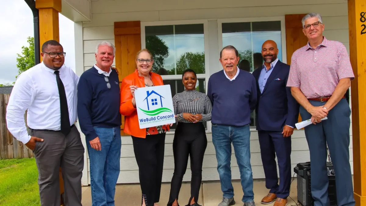 Seven people standing in front of a house, one holding a "We Build Concord" sign.