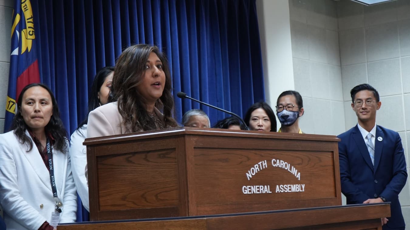 Woman speaking at a North Carolina General Assembly podium with six people standing behind her.