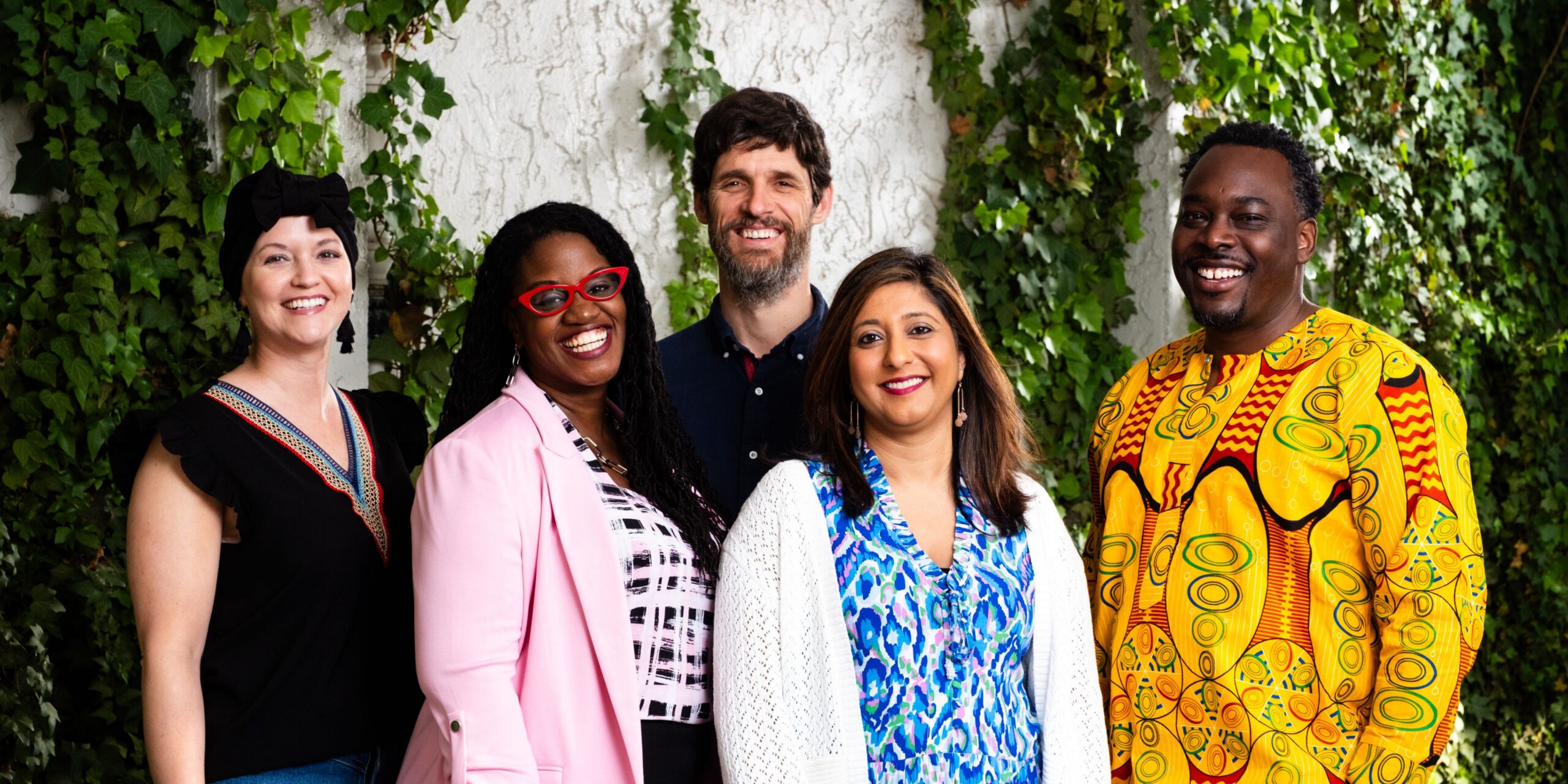 Five smiling adults posing together in front of an ivy-covered wall.