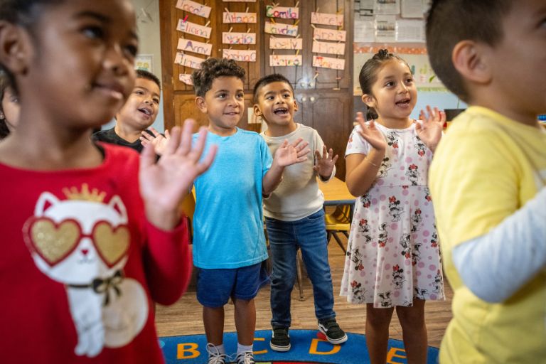 Five young children stand in a preschool classroom, smiling and clapping