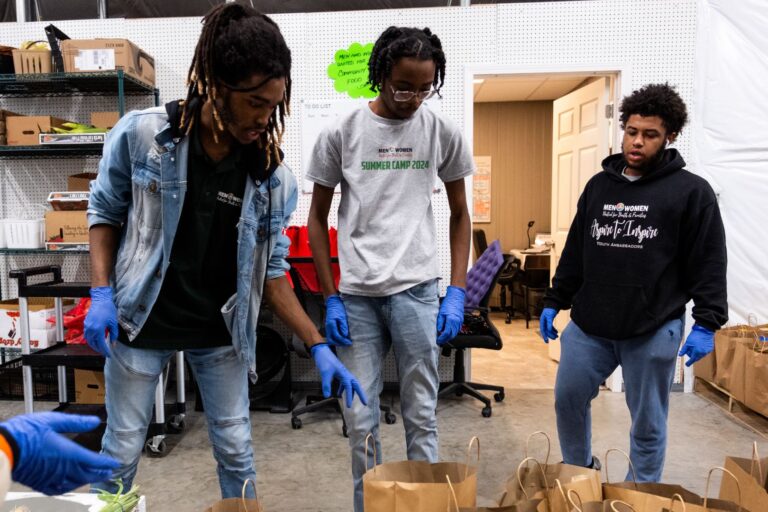 Three young men stand in front of empty produce bags in a warehouse