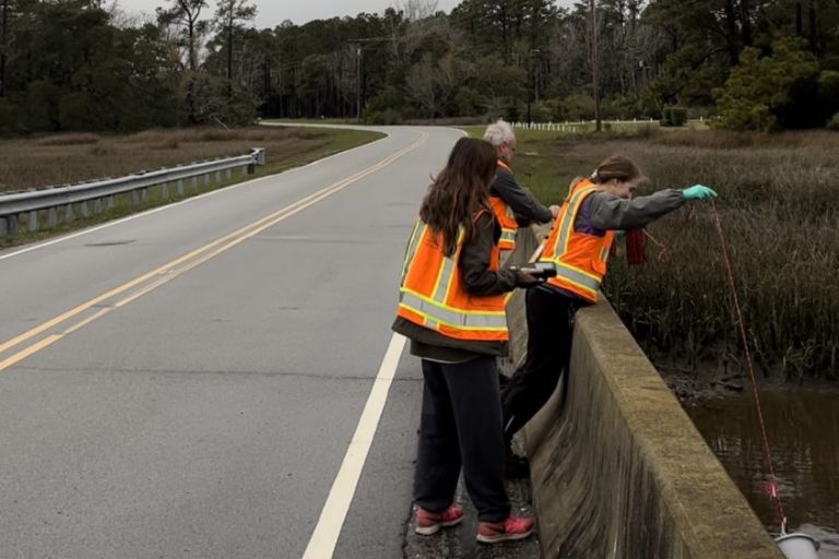 Three researchers lean over the edge of a bridge to test water quality in the river below.