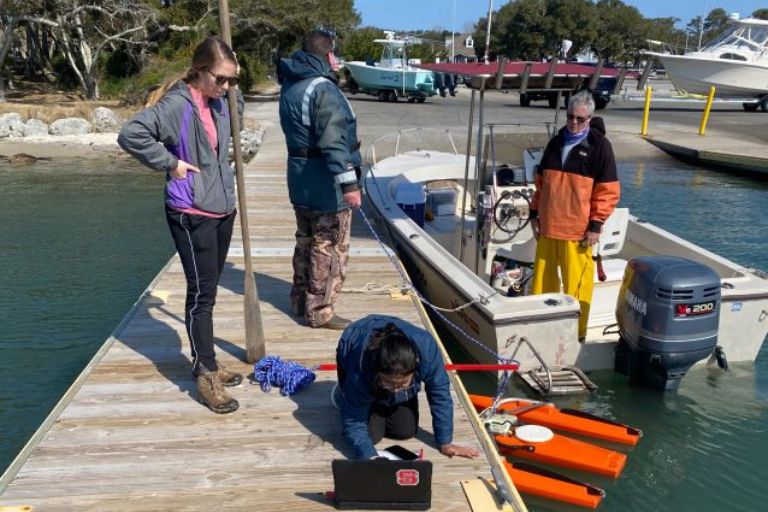 Three researchers stand on a dock preparing to collect water samples. A man stands in a boat next to the dock.