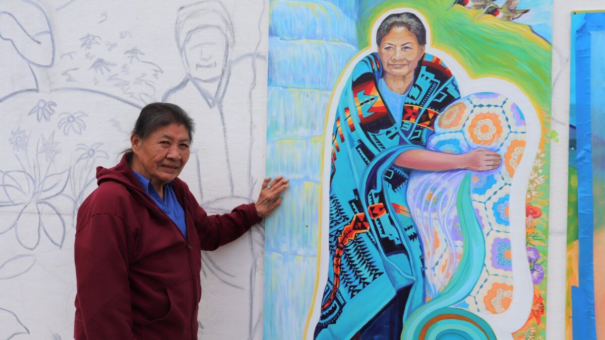 Ella Bird, an elderly woman from the Snowbird Cherokee community, stands next to a mural with her portrait painted on it