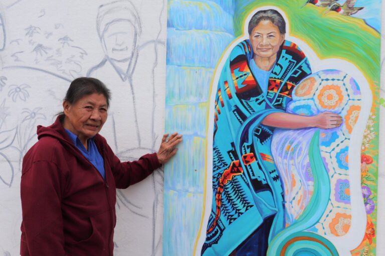 Ella Bird, an elderly woman from the Snowbird Cherokee community, stands next to a mural with her portrait painted on it