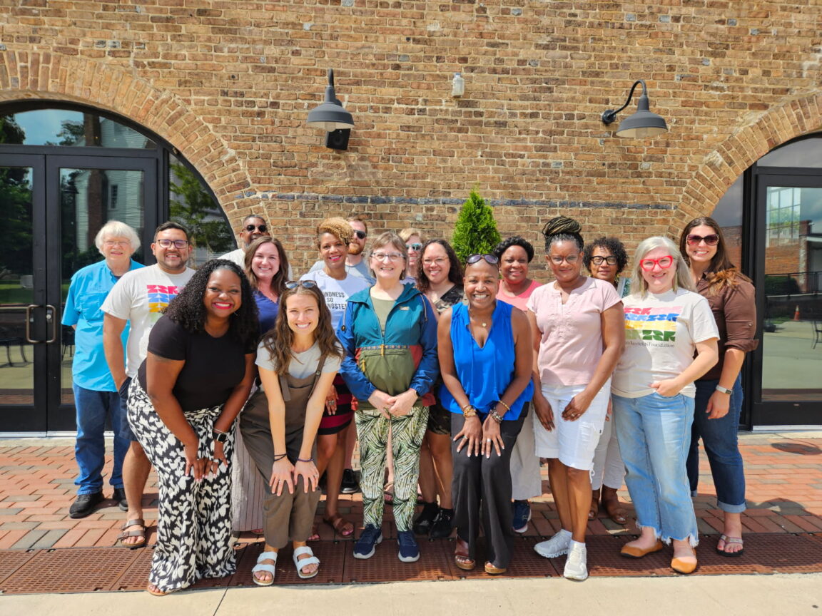 Group of 17 diverse adults smiling outdoors in front of a brick building with arched windows and doors.