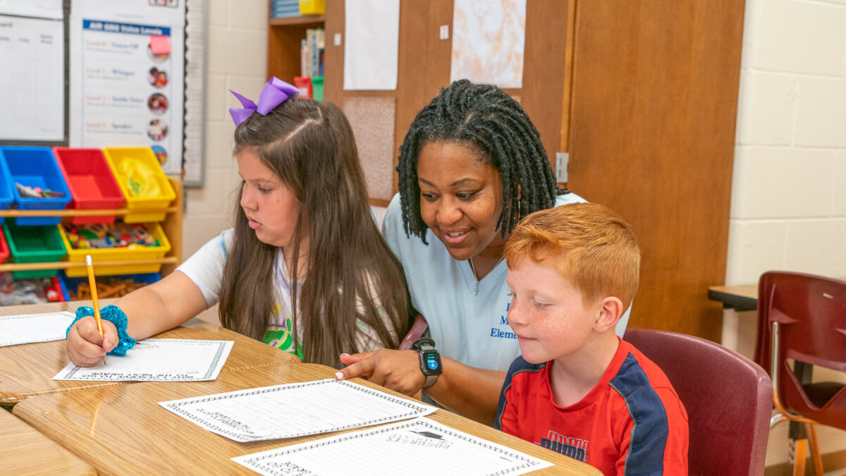 Teacher helping two elementary students with writing assignments at a classroom desk