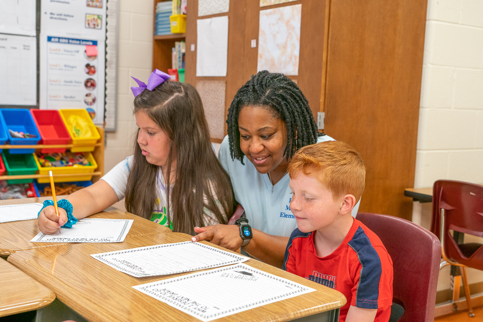 Teacher helping two elementary students with writing assignments at a classroom desk