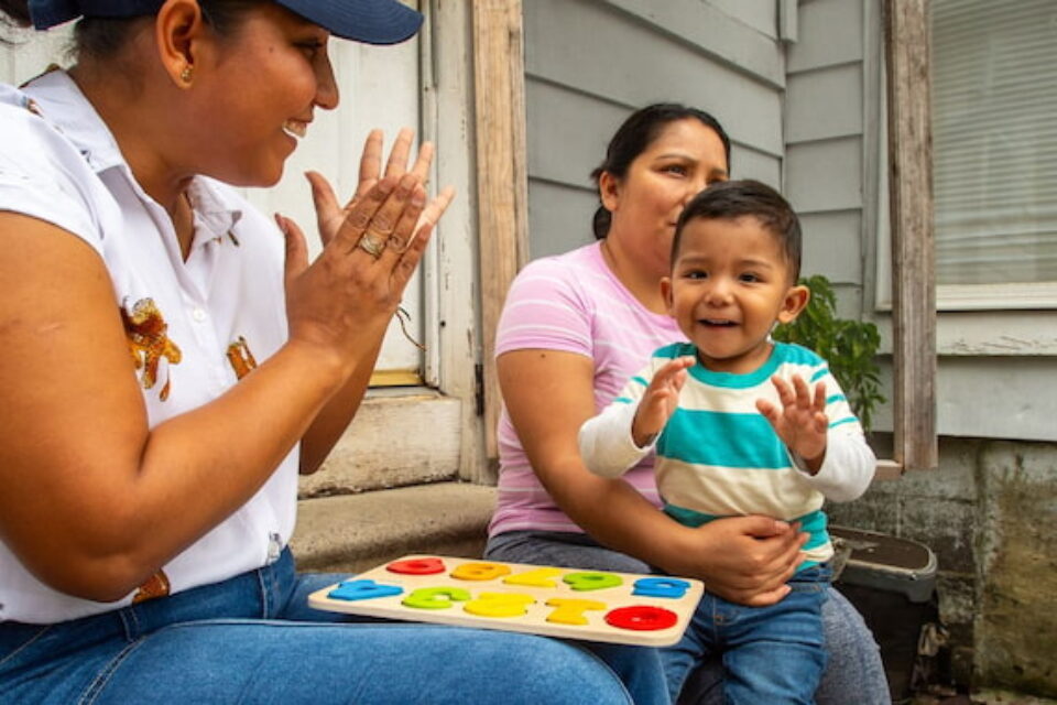 Woman clapping hands while a toddler on another woman's lap plays with a colorful number puzzle outdoors.