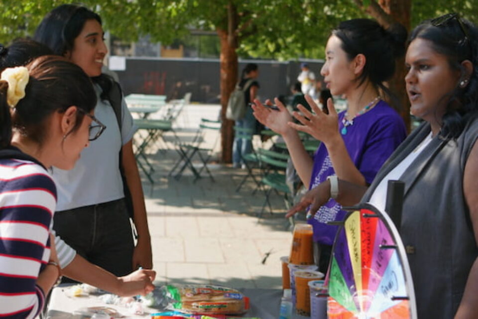 Four women engaged in conversation at an outdoor table with a colorful prize wheel and drinks.
