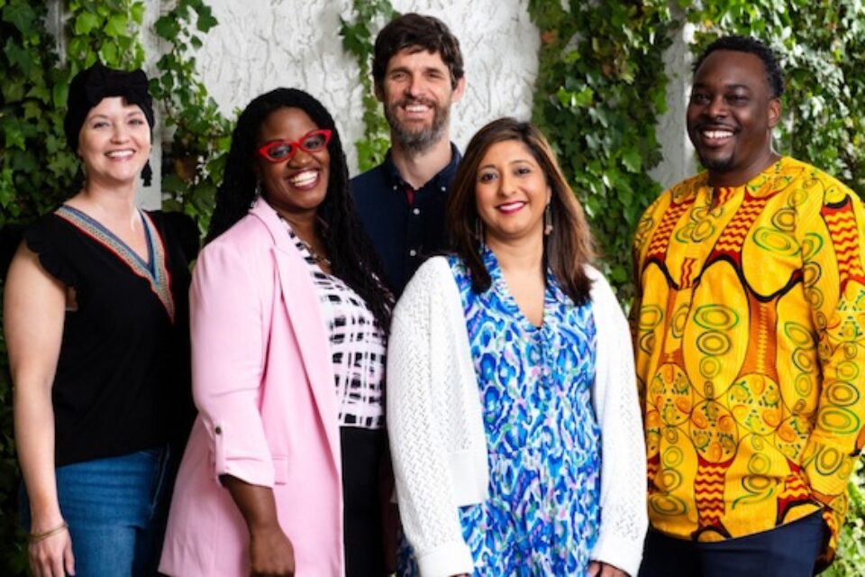 Group of five diverse adults smiling, standing outdoors with green ivy on a white wall background.