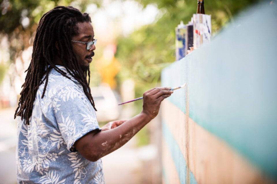 Man with dreadlocks painting a blue and beige wall outdoors with a paintbrush.
