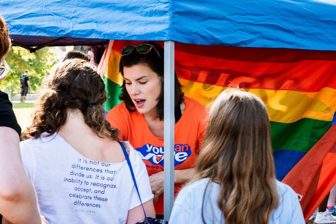 People gathered under a rainbow Pride flag tent, talking at an outdoor event.