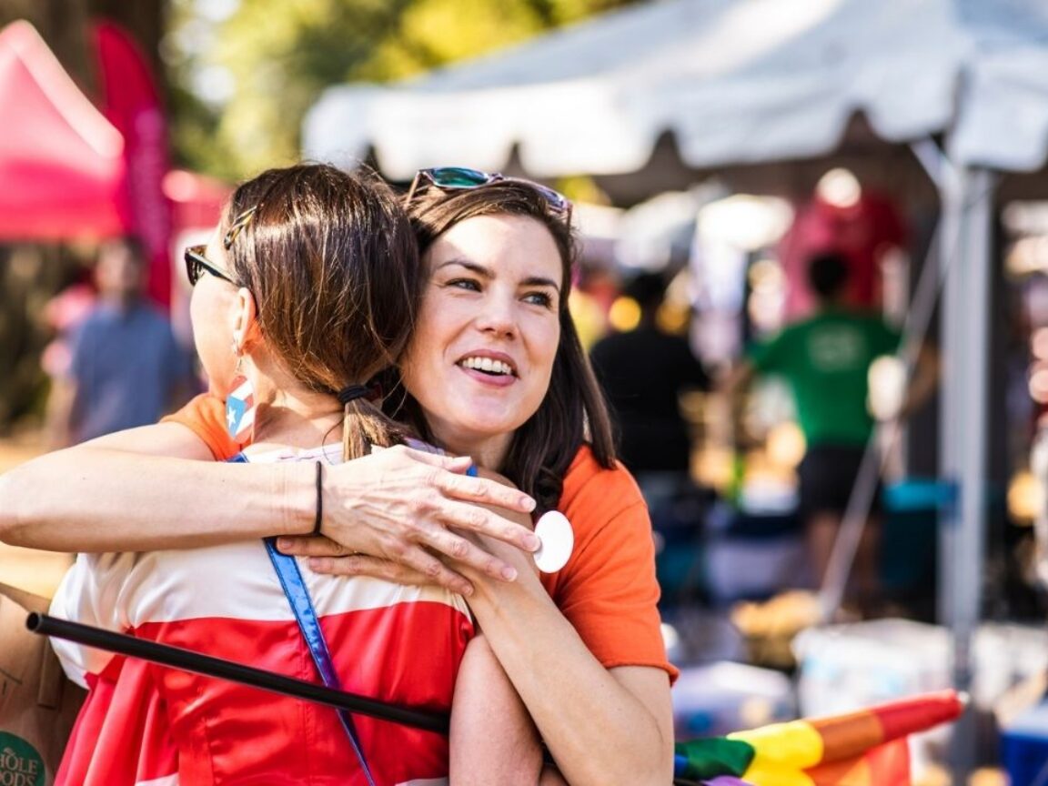 Two women hugging at an outdoor festival with tents and crowds in the background