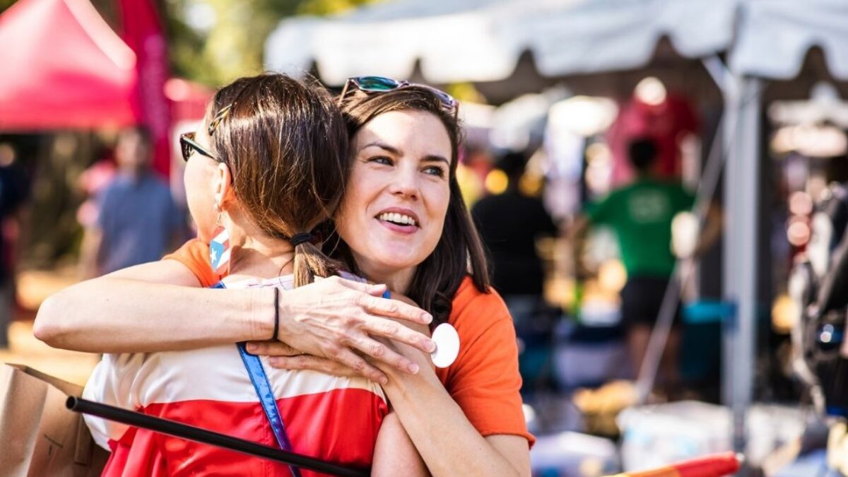 Two women hugging at an outdoor festival with tents and crowds in the background