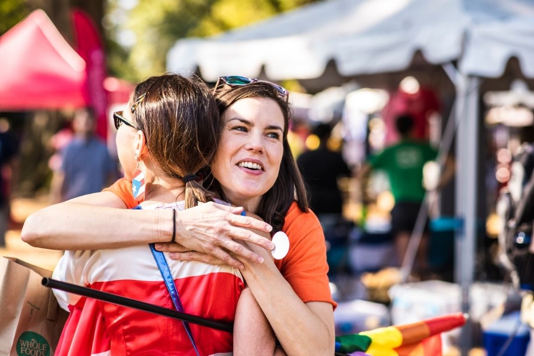 Two women hugging at an outdoor festival with tents and crowds in the background