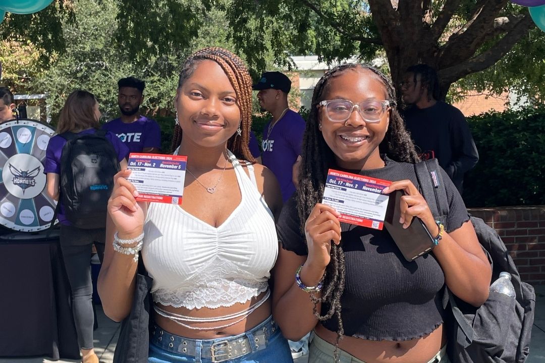 Two smiling women holding North Carolina elections information cards at an outdoor campus event.