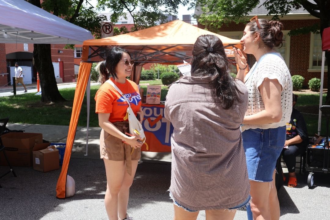 People chatting at an outdoor booth under an orange canopy at a community event