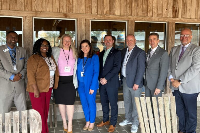 Seven adults in business attire posing together outside a wooden building.