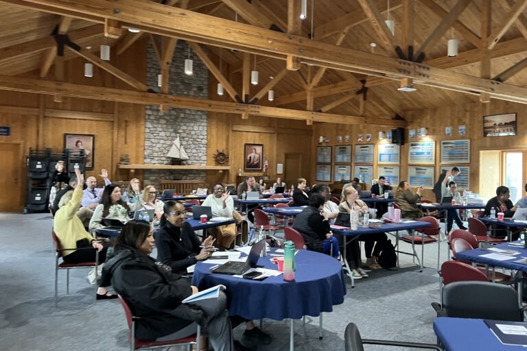 Workshop attendees seated at tables in a wood-paneled hall, many raising hands during a group session.