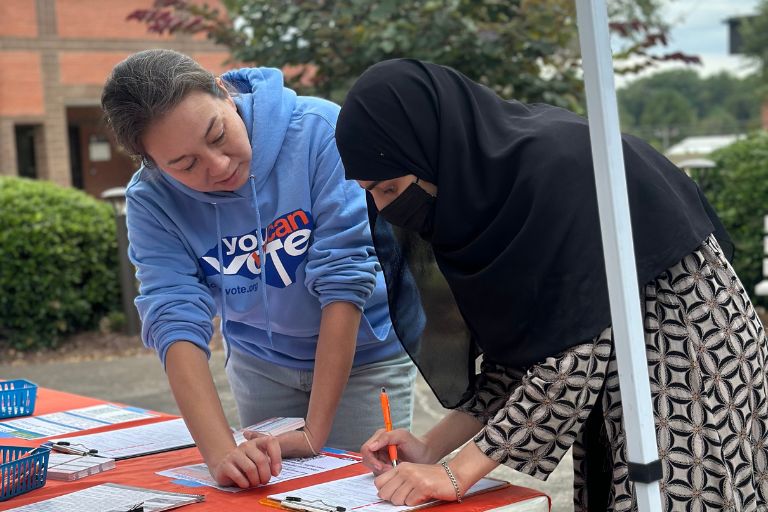 Two women fill out forms at an outdoor voter registration table.