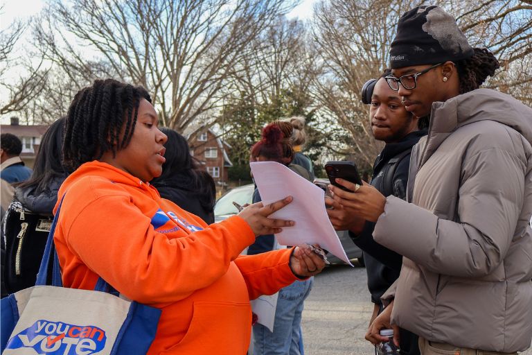 Person in orange hoodie hands out papers to two people checking a phone outdoors