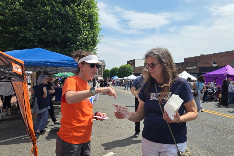Two women exchanging flyers at an outdoor street fair with vendor tents and crowds
