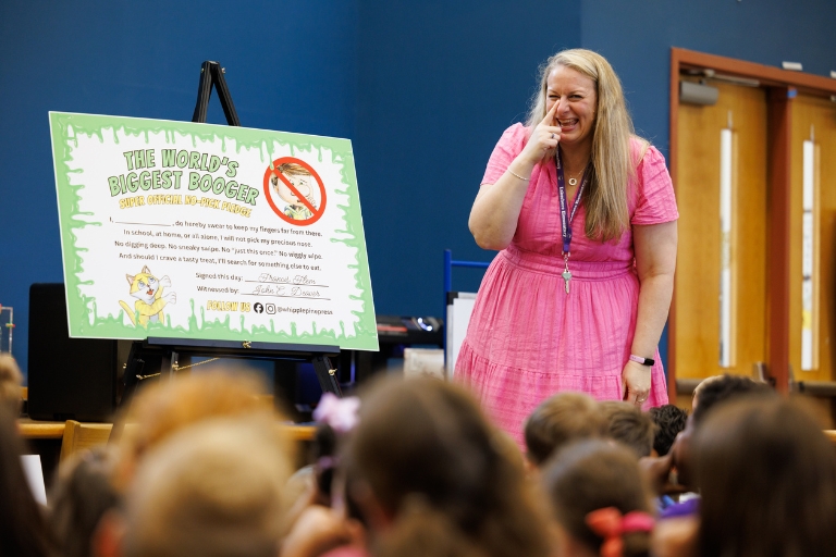 Principal beside poster titled “The World’s Biggest Booger” speaking to seated children