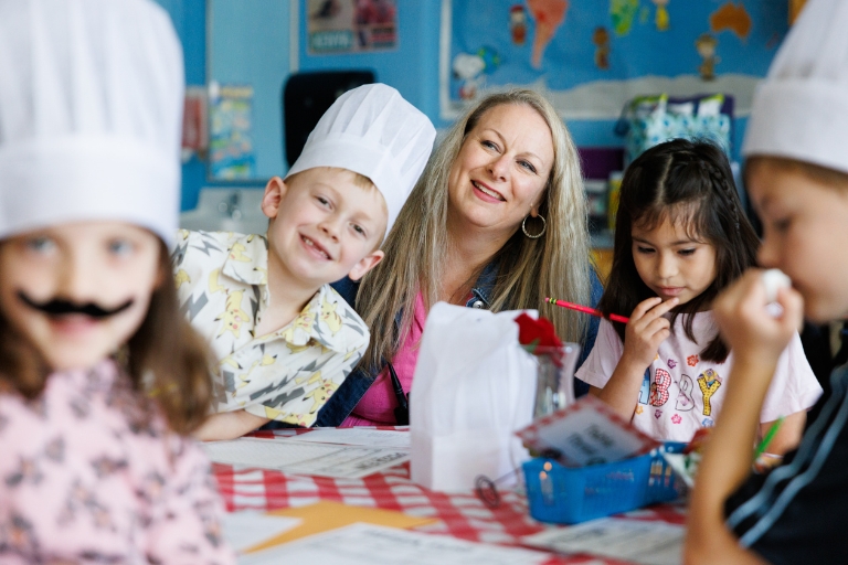 Principal with children in a classroom, some wearing chef hats, gathered around a table doing an activity.