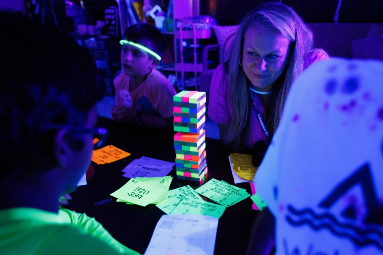 Children and a principal play a neon-lit block-stacking game at a table with score cards.