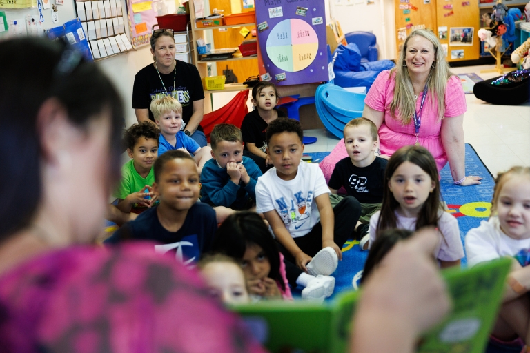 Principal reading to a group of young children seated on a classroom rug with two adults nearby
