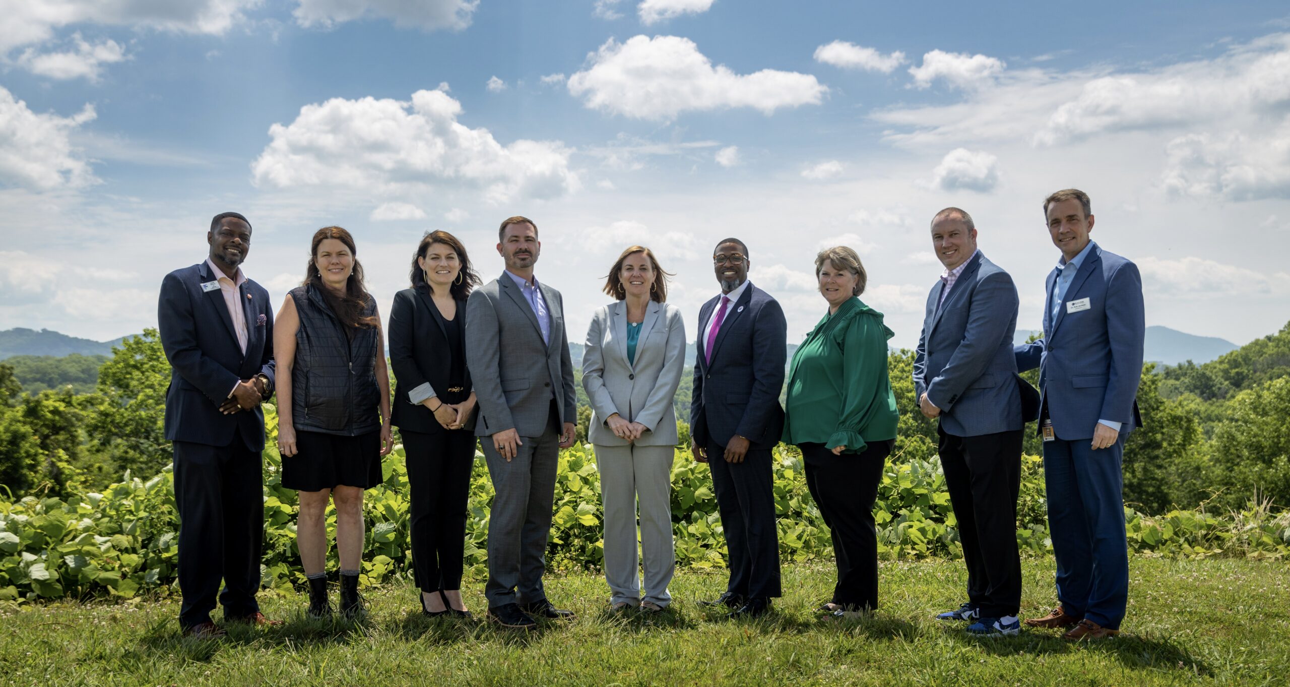 Eight people in business attire standing outdoors on a grassy hill under a cloudy blue sky.
