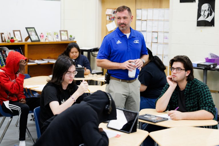 Principal stands among students in a classroom, holding a cup while students work at desks.