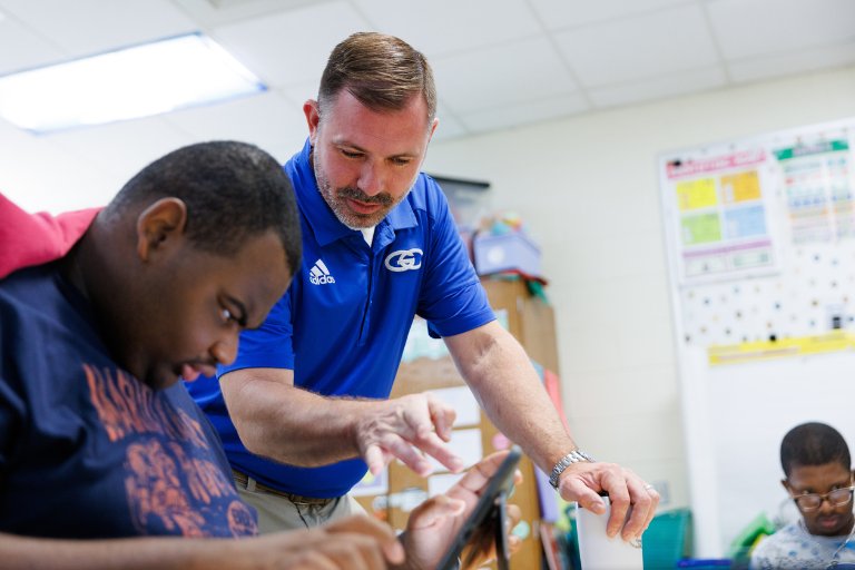 Principal helping a student use a tablet in a classroom while another student watches nearby