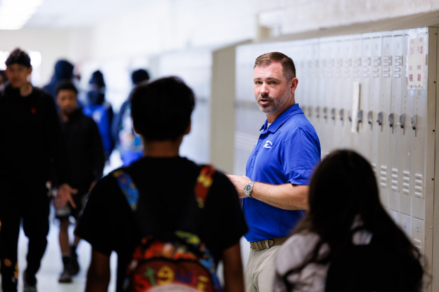 Teacher in blue polo speaking to students in a school hallway beside lockers